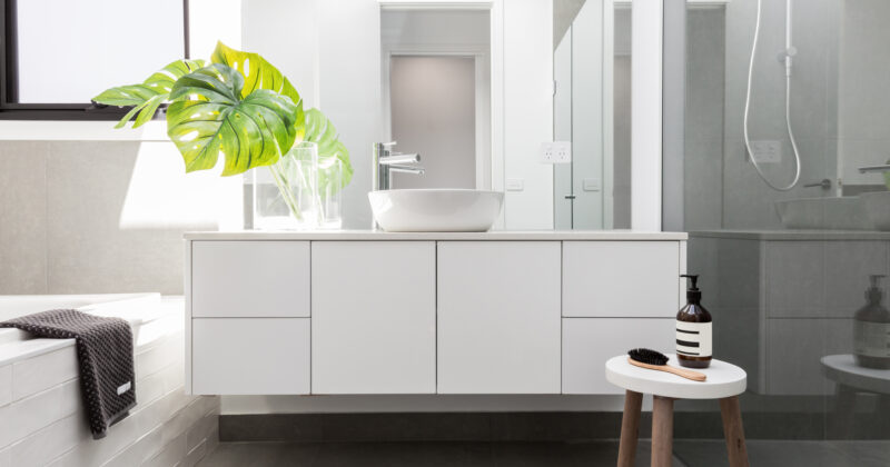 Modern bathroom with a white vanity, vessel sink, and large mirror. A glass shower enclosure is on the right, a potted plant on the left, and a small stool with a soap dispenser and brush in the foreground. Explore our website for the best bathroom cabinets to complete your design.