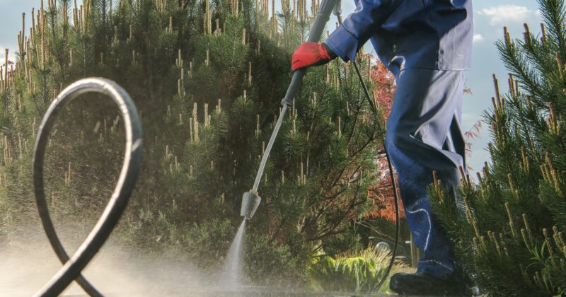 A person in blue overalls and red gloves is pressure-washing a concrete surface outdoors, with green trees in the background.