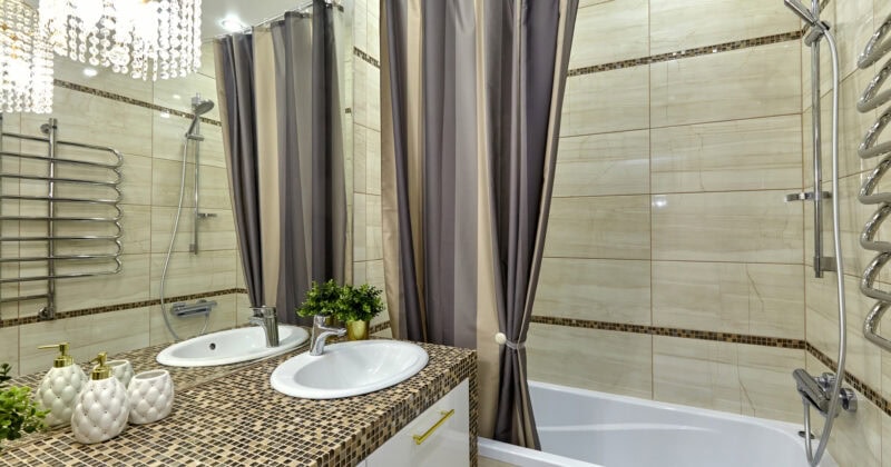 A modern bathroom remodel with a bathtub, shower, and two sinks. Beige tiled walls and countertops, striped shower curtains, and a chandelier-style light fixture complete the decor—a perfect showcase for Bathroom Remodelers' expert touch.