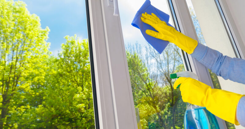 A person in yellow gloves is expertly engaged in window cleaning, using a blue cloth and spray bottle. Through the spotless glass, trees and a clear blue sky are visible, showcasing the best way to brighten up any view.