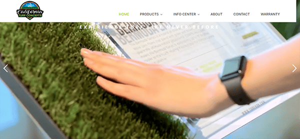 A hand with a smartwatch touches artificial grass mounted on a display stand. The background shows a blurred brochure and parts of a website header with navigation links.