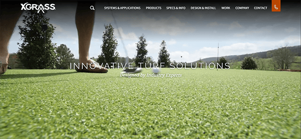 Close-up view of artificial turf with a person's legs and feet in sandals visible in the foreground; background features trees and hills. Text on image reads "Innovative Turf Solutions, Designed by Industry Experts.