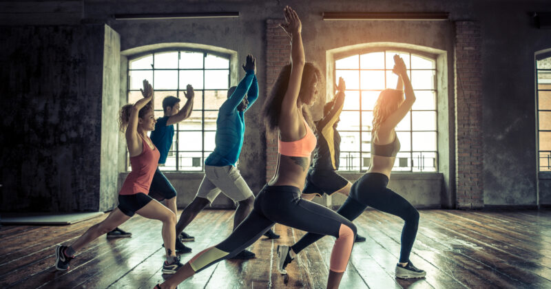 A group of people in athletic wear exercise in a sunlit studio, performing lunges with their arms raised. The room, reminiscent of spaces featured in the best fitness website designs, boasts large windows and wooden floors.