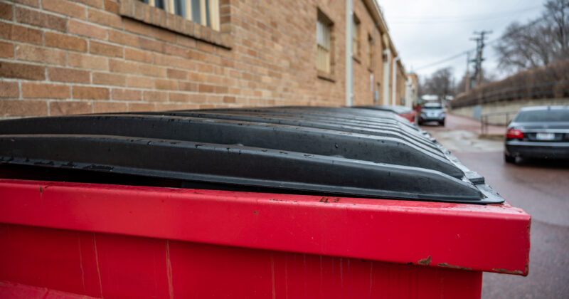 A red dumpster with a closed black lid is positioned beside a brick building on a wet street, evoking the clean and user-friendly feel of a well-designed dumpster rental website, with parked cars in the background.