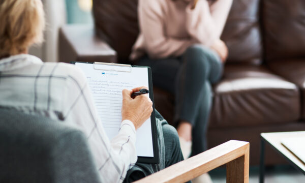 Two people in a counseling session: a therapist from a private practice taking notes on a clipboard and a client sitting on a brown sofa.