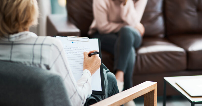 Two people in a counseling session: a therapist from a private practice taking notes on a clipboard and a client sitting on a brown sofa.