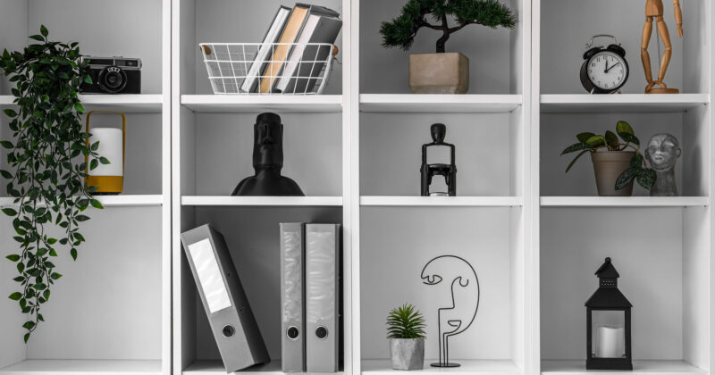 A white shelving unit displaying various items, including books, binders, plants, a camera, a clock, and artistic décor—perfect for those seeking the best home organizing solutions.