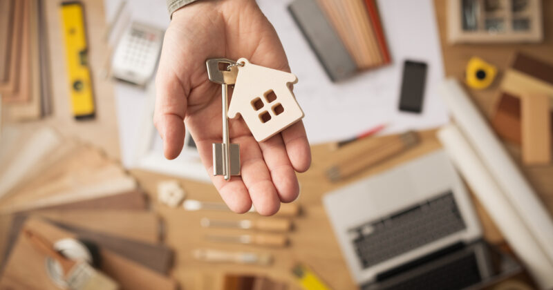A person's hand holds a house key with a wooden house-shaped keychain. The table below, reminiscent of a busy real estate office, features various tools, wood samples, a laptop displaying website designs, and papers scattered about.