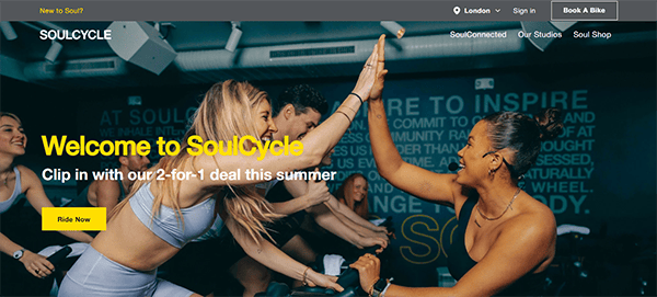 Group of people in an indoor cycling class high-fiving each other. Text on image advertises a 2-for-1 deal at SoulCycle. "Welcome to SoulCycle" and "Ride Now" buttons are present.