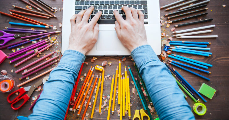 Hands typing on a laptop surrounded by scattered colored pencils, pens, scissors, and other stationery items on a wooden surface — the perfect setup for crafting stunning Portfolio Websites or starting your next Graphic Design masterpiece.