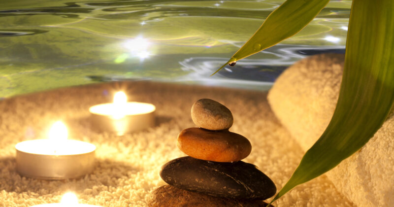 A stack of four stones on a white towel, accompanied by three lit tealight candles and a green leaf with water droplets, is set against a serene water backdrop perfect for wellness websites.