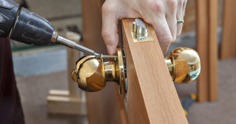 A person uses a power drill to install a gold-colored doorknob on a wooden door, having consulted the best locksmith websites for expert advice.