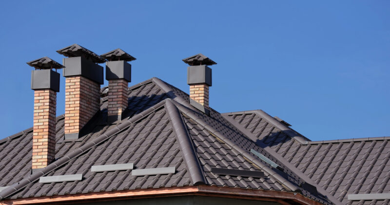 A close-up of a residential roof with dark shingles and multiple brick chimneys against a clear blue sky, perfect for showcasing the craftsmanship advertised on chimney repair websites.