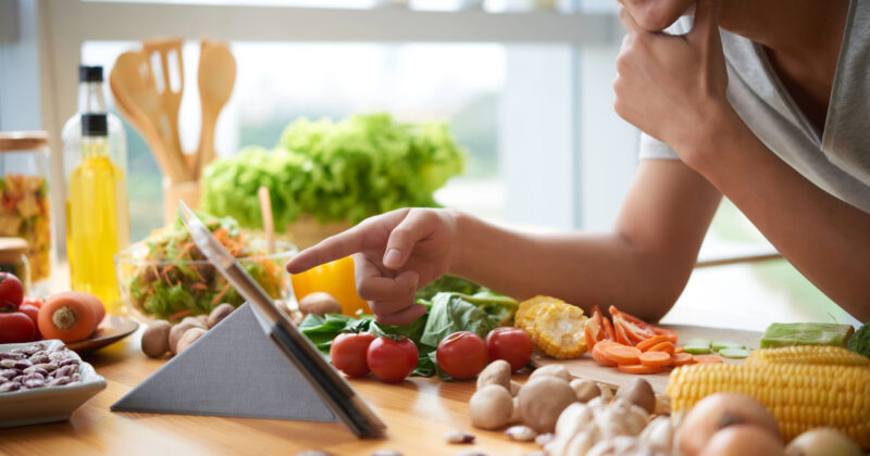 A person uses a tablet while surrounded by various fresh vegetables in a kitchen, browsing through the best recipe websites to find culinary inspiration.