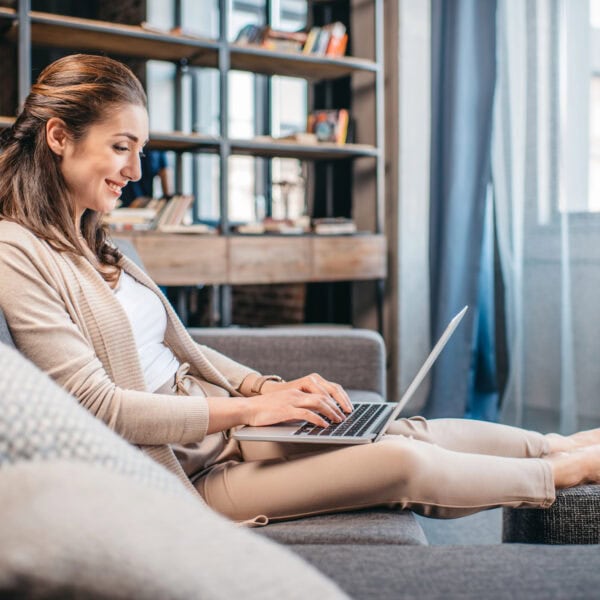 A woman sitting on a gray sofa, working on a laptop in a modern living room with shelves and books in the background, strategically planning her online marketing strategies.