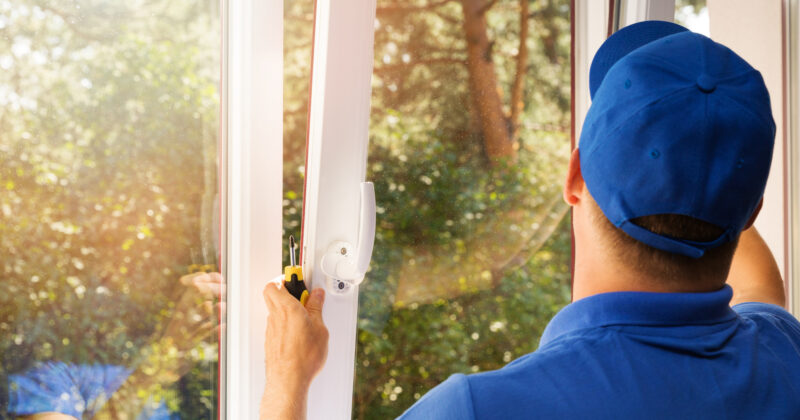 A man in a blue uniform installs a white-framed window, holding a screwdriver with trees visible outside. For more information on his services, check out the best window repair websites.