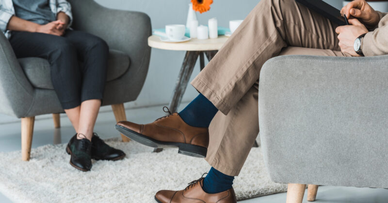 Two individuals sitting in a counseling session. One, likely the psychotherapist, has crossed legs and holds a notepad, while the other is seated with visible shoes. A small table with a flower and cups sits in the background. This cozy setting could be featured on the best websites for mental health resources.