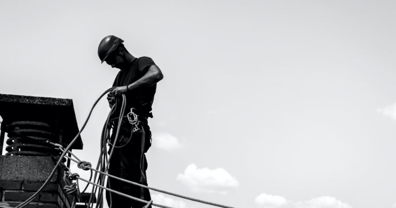 A worker in a safety helmet and harness handles ropes while standing on a rooftop near a chimney, performing chimney cleaning against a clear sky.