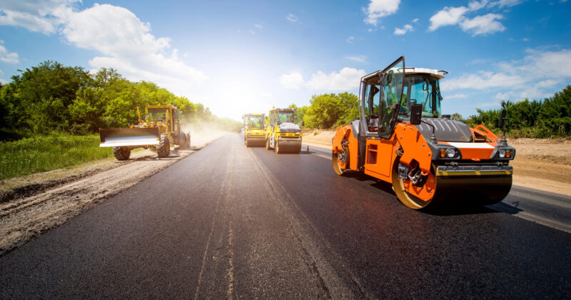 Construction vehicles paving a new road under a clear blue sky, including a road roller in the foreground and other machinery working in the background, provide an ideal scene for showcasing asphalt sealcoating techniques.