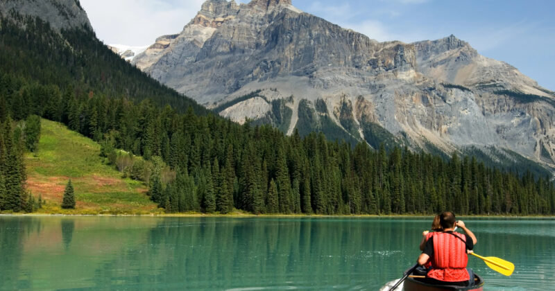 Person paddling a red canoe on a calm, turquoise lake surrounded by dense forests and towering mountains under a clear sky, ideal for those exploring the best tourism websites for travel inspiration.