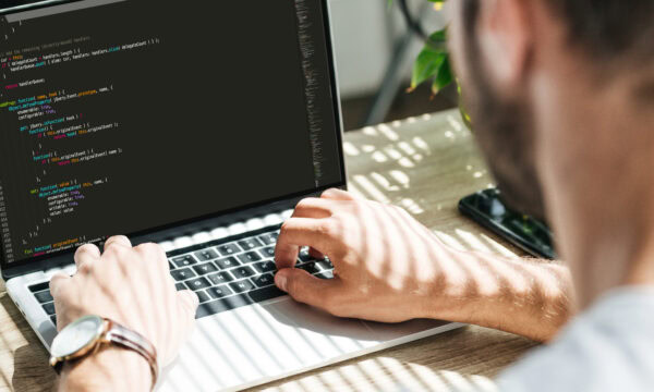 A person is typing code on a laptop with a black screen displaying programming syntax. Taken over the shoulder, natural light filters through, highlighting the plant in the background. As they work on the new website cost estimates for 2024, they focus intently on every detail.