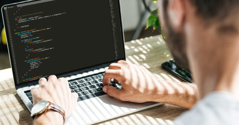 A person is typing code on a laptop with a black screen displaying programming syntax. Taken over the shoulder, natural light filters through, highlighting the plant in the background. As they work on the new website cost estimates for 2024, they focus intently on every detail.