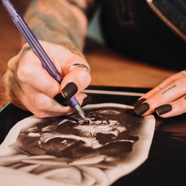 A person with tattoos and painted nails, likely a tattoo artist, is using a purple pen to work on a grayscale image of a human skull.