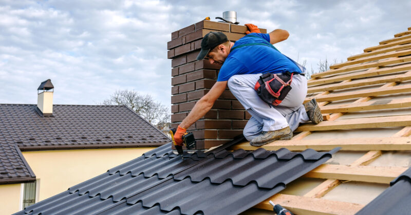 A worker in a blue shirt and white pants installs black roof tiles on a wooden roof structure near a brick chimney under a cloudy sky, showcasing the quality of residential roofing.