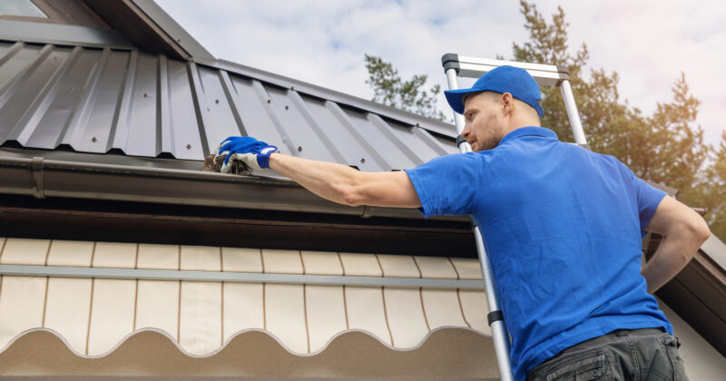 A person in a blue shirt and cap stands on a ladder cleaning debris from a house gutter. The house, similar to those featured on the best websites for industrial electrician housing, has a metal roof and awning.