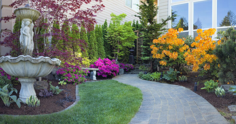 A well-maintained garden with a stone pathway, vibrant flowers, a tall fountain with a statue, and various greenery adorns the house with large windows—an inspiration for those seeking hardscape design.