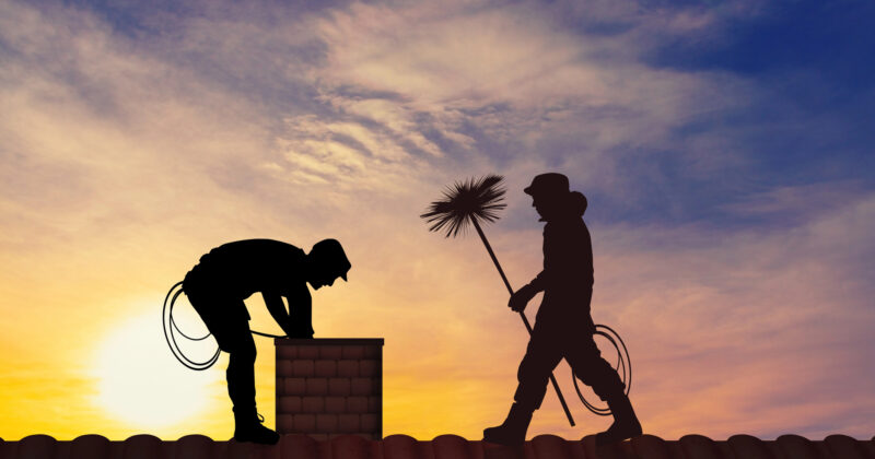 Silhouettes of two workers on a rooftop at sunset, one bending over a chimney and the other holding a chimney sweep tool—classic professionals from the best chimney sweep websites.