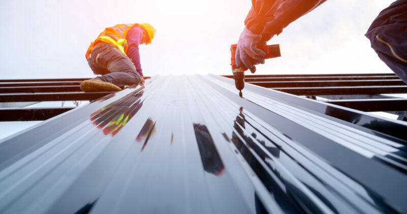 Two construction workers from one of the best roofing websites are installing a reflective metal roof panel, with one using a power drill. Both are wearing safety gear, including helmets and high-visibility vests.