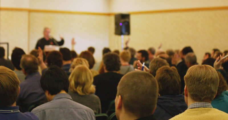 A person at the front of a crowded room gives a speech while the seated audience raises their hands, illustrating the warmth and engagement you can find at Best Church.