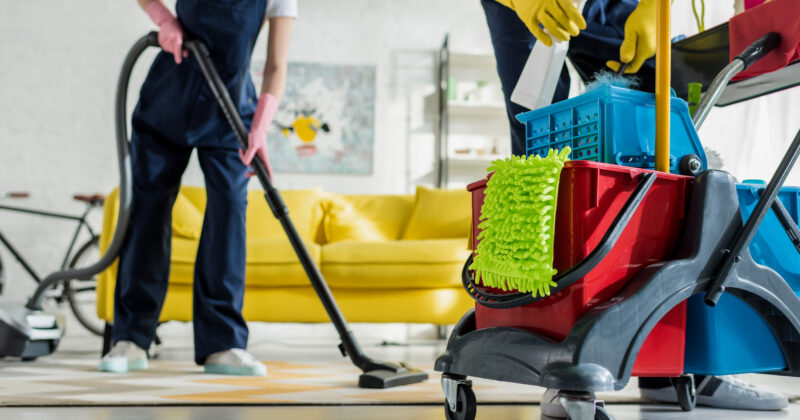 Two uniformed individuals are meticulously cleaning a living room. One handles the vacuum cleaner, focusing on carpet cleaning, while the other manages cleaning supplies on a cart. The room features a yellow couch and various visible tools, capturing the essence of top services one might find on the best websites.