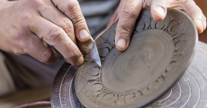 Close-up of hands carving intricate designs into a ceramic plate using a small carving tool. The plate is held on a rotating platform. Discover more about this craft on the best artisan websites, where masters share their techniques.