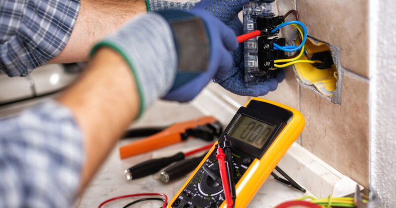 An electrician's hands using a multimeter to test electrical wiring in a wall socket, surrounded by tools and wires, showcasing the skills often featured on residential electrician websites.