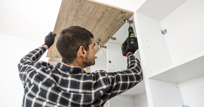 A person wearing gloves and a plaid shirt uses a power drill to assemble white furniture, attaching a wooden panel to the top—a scene typical of the best cabinet makers showcased on cabinet maker websites.