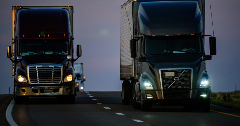 Two semi-trucks drive on a highway at dusk, their headlights cutting through the dimly lit sky, which is a gradient of purple and blue. One truck occupies the left lane, while the other stays in the right. For more insights and resources on trucking, check out 20 Best Trucking Websites available online.