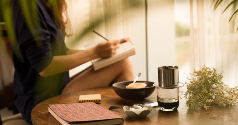 A person sits by a window writing in a notebook, inspired by ideas for the best author websites. A table in front holds a journal, coffee setup, and a bowl of food. White flowers and greenery partially frame the view.