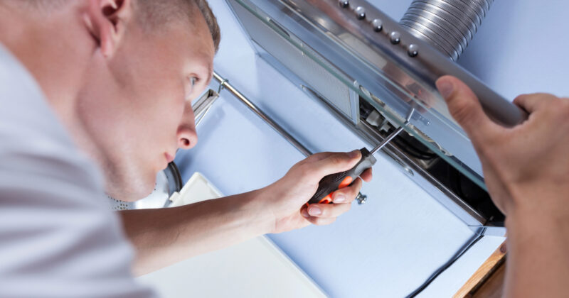 A person is using a screwdriver to perform appliance repair on a metallic device in the kitchen, drawing on tips from the best appliance repair websites.