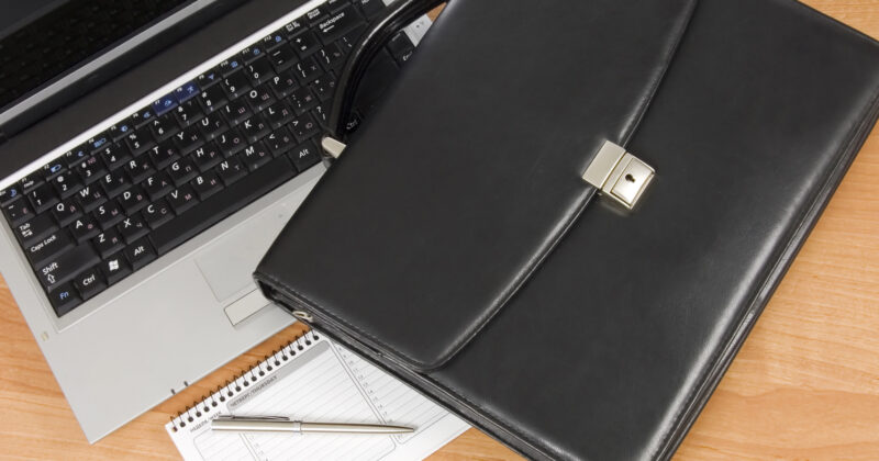 A black briefcase, a silver laptop displaying 20 Best Personal Portfolio Websites, a lined notepad, and a pen are placed on a wooden desk.