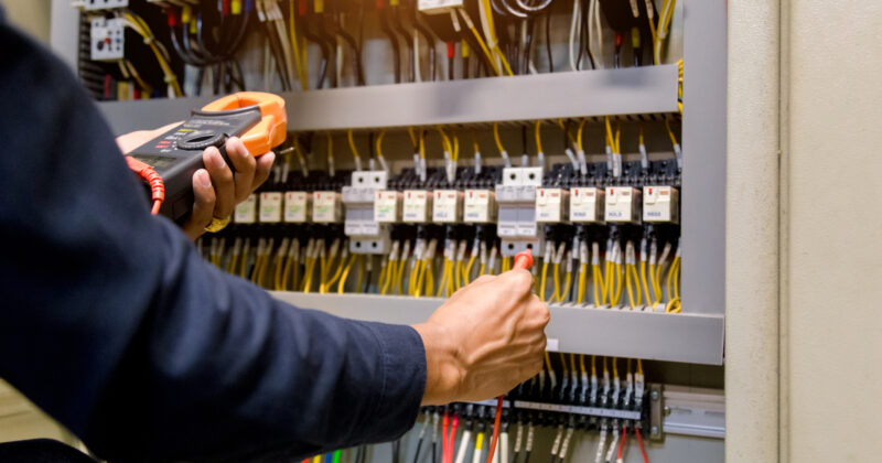 A person is using a multimeter to check electrical connections inside a circuit breaker panel. The panel is filled with wires and various electrical components, showcasing the expertise found on top Commercial Electrician Websites.