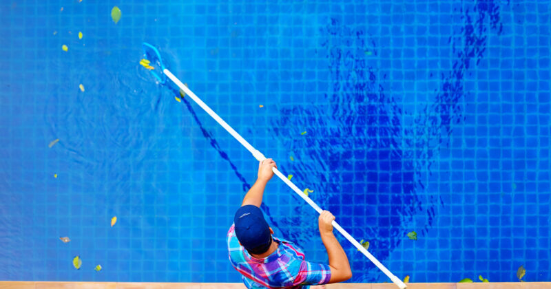 A person wearing a plaid shirt and cap uses a long pool skimmer to remove leaves from a blue-tiled swimming pool, likely inspired by tips from the best pool cleaning websites.