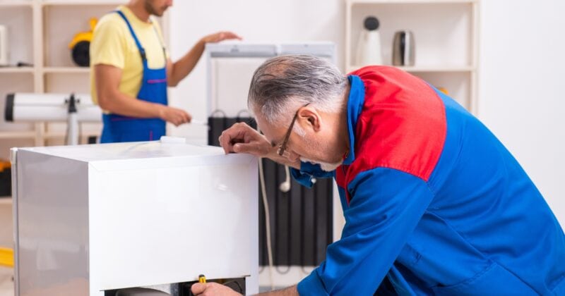 Two technicians in a workshop—one in the foreground focused on refrigerator repair with tools sprawled on the table, while another works on a different appliance in the background.