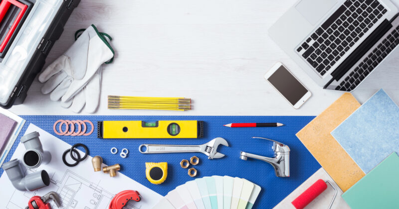 Various tools and materials for home improvement projects are arranged on a work surface, including a laptop displaying the best home services websites, a smartphone, blueprints, pliers, a wrench, gloves, color swatches, and measuring tape.