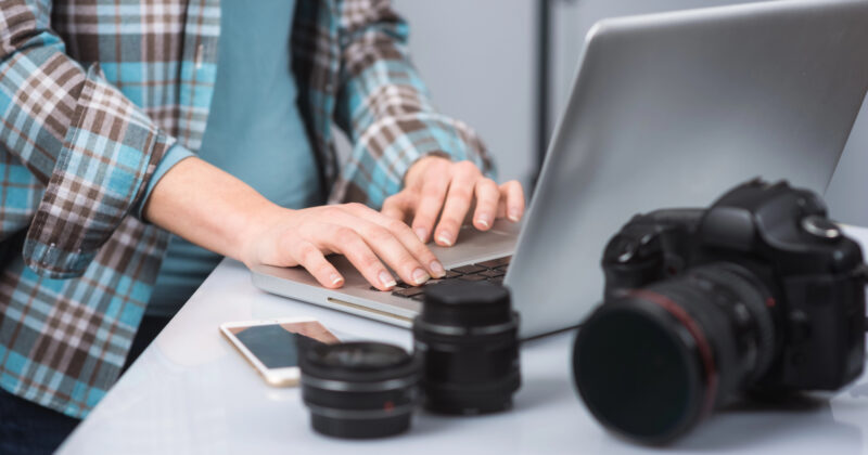 A person typing on a laptop at a desk surrounded by camera lenses, a camera, and a smartphone, possibly researching the best photography platforms.