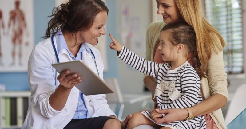 A female pediatrician with a tablet smiles at a young girl sitting on a woman's lap in the medical office. The child is pointing excitedly at the doctor.