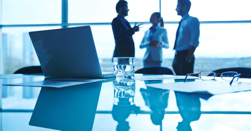 A glass of water, a pair of glasses, and documents on a table with a laptop, as three people discuss the best websites for small business in the background.