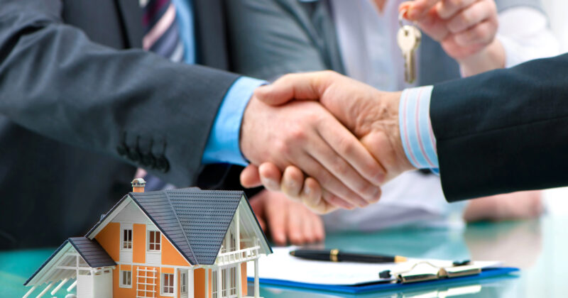 A close-up of two people shaking hands over a desk with a model house, pen, clipboard, and another person handing over keys in the background—a scene often featured on the 20 best realtor websites.