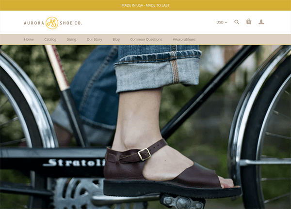 Close-up shot of a person's foot on a bicycle pedal, wearing dark brown sandals. The background has blurred parts of the bicycle and denim pants. Aurora Shoe Co. logo and website navigation bar are visible at the top.